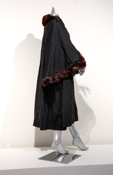 Photograph taken from the side of a black ceremonial robe with brown fur trim on a silver mannequin.