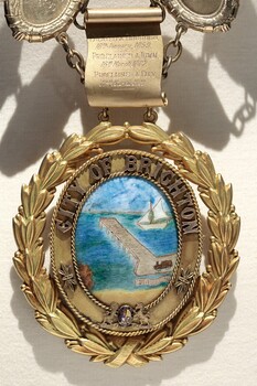 Photograph of the Oval drop medallion of the City of Brighton seal illustrating a coastline with a pier extending into the water and a sailboat in the distance.