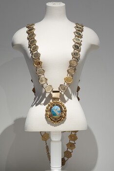 Photograph of a gold ceremonial Mayoral chain on a cream torso. The chain consist of small oval links, with a large drop medallion of the City of Brighton seal.