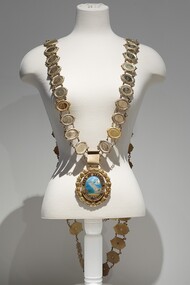 Photograph of a gold ceremonial Mayoral chain on a cream torso. The chain consist of small oval links, with a large drop medallion of the City of Brighton seal.