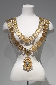 Photograph of a gold ceremonial mayoral chain consisting of small oval links and a large drop medallion on a mannequin torso.