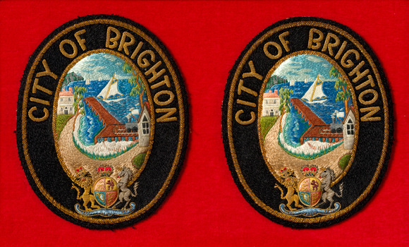 A pair of oval embroidered City of Brighton Mayoral Badges on a red felt background. The seal has a black border and depicts a coastline with a pier, sail boats, train, and buildings. Below it is the British Coat of Arms.