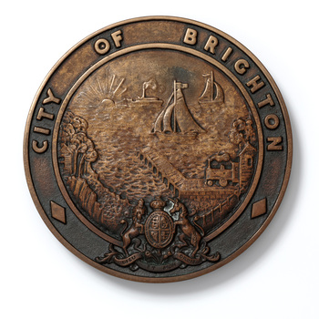 Round cast bronze plaque for City of Brighton crest, with a coastal scene in the centre and British Coat of Arms at the bottom.