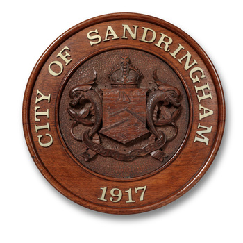 Round wooden carved relief plaque with the metal lettering around the boarder 'CITY OF SANDRINGHAM 1917' and a coat of arms in the centre featuring a shield flanked by 2 dolphins 
