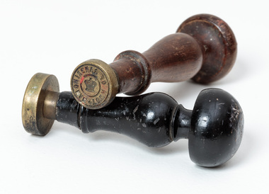 Photograph a brown handled seal sitting on a black handled seal. The brass seal has writing around the edge and a crown at centre