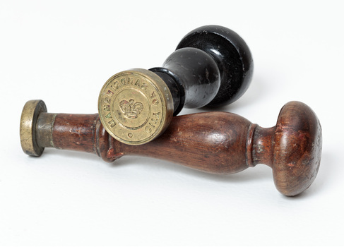 Photograph a black handled seal sitting on a brown handled seal. The brass seal has writing around the edge and a crown at centre