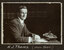 Black and white photograph of a man in a suit seated at a desk with his name written underneath on the mount