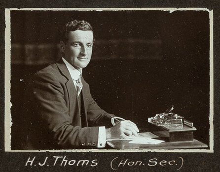 Black and white photograph of a man in a suit seated at a desk with his name written underneath on the mount