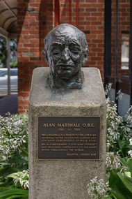 Cast bronze head of a balding man on a cement pedestal with bronze plaque at front, situated in a garden outside a building