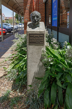Cast bronze head of a balding man on a cement pedestal with bronze plaque at front, situated in a garden outside a building