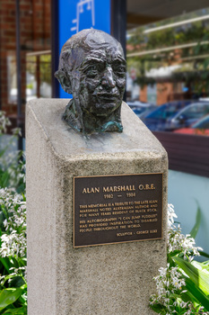Cast bronze head of a balding man on a cement pedestal with bronze plaque at front, situated in a garden outside a building