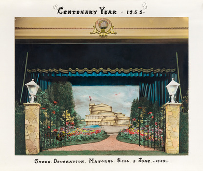 A hand-coloured photograph of a stage with dark curtains open to a set scene of the new Brighton Municipal Centre flanked by gardens.