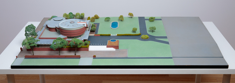 Architectural model of a cylindrical building in orange brick surrounded by gardens and paths