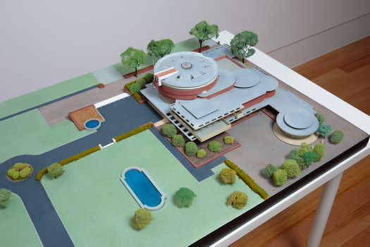 Architectural model of a cylindrical building in orange brick surrounded by gardens and paths, photographed from above