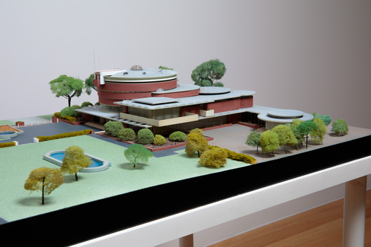 Architectural model of a cylindrical building in orange brick surrounded by gardens and paths
