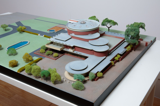 Architectural model of a cylindrical building in orange brick surrounded by gardens and paths