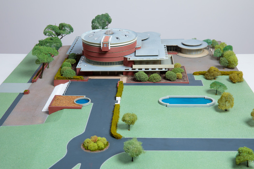 Architectural model of a cylindrical building in orange brick surrounded by gardens and paths