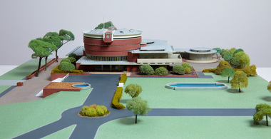 Architectural model of a cylindrical building in orange brick surrounded by gardens and paths