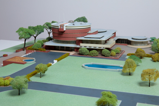 Architectural model of a cylindrical building in orange brick surrounded by gardens and paths