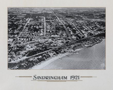 Aerial black and white photograph of a coastal suburb, taken from over the bay with the beach in the foreground and grids of house lined streets in the background. 'SANDRINGHAM 1921' written below image