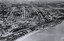Black and white aerial photograph of a coastal suburb, taken from over the bay, with the beach in the foreground and grids of house-lined streets in the background.