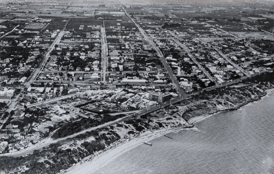 Black and white aerial photograph of a coastal suburb, taken from over the bay, with the beach in the foreground and grids of house-lined streets in the background.