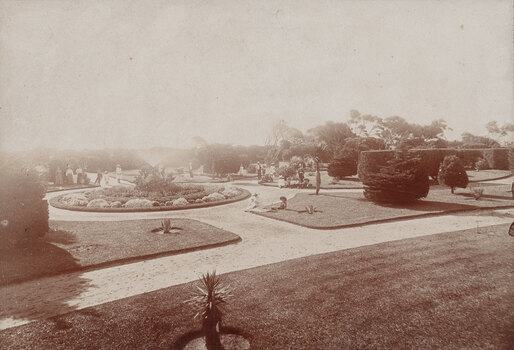 Sepia toned photograph taken from a high vantage point of a formal garden with winding paths and vegetation. A group of people in early 20th century clothes are posing for the camera