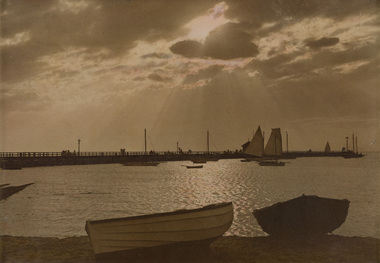 Sepia toned photograph of a coastal scene with a large pier in the middle distance, water and boats on the shore, the sun's rays appearing out through the clouds above. 