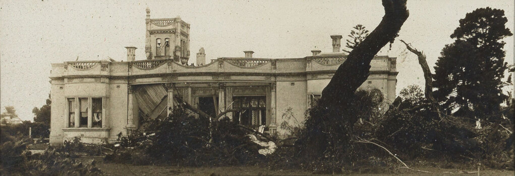 Sepia photograph of a grand house with a tower that has been damaged by cyclone - the verandah has collapsed, and trees are broken in the garden.