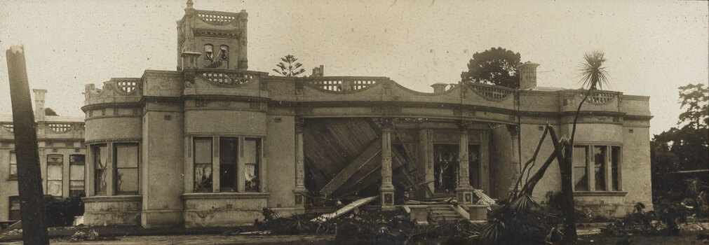 Sepia photograph of a grand house with a tower that has been damaged by cyclone - the roof of the front verandah has collapsed, and trees are broken in the garden