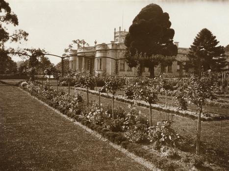 Black and white photograph of a grand house in the distance with lawns and rose garden beds in the foregraound