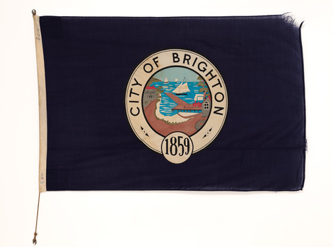Navy blue flag for City of Brighton with frayed corner. In the centre is the crest showing the coastline, a pier, train and sail boats.
