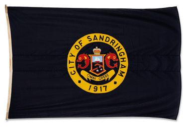 Dark blue flag with City of Sandringham crest in centre. The crest depicts a shield with a boat and pier scene, flanked by 2 red dolphins and the St Edward's crown above. A motto in latin below