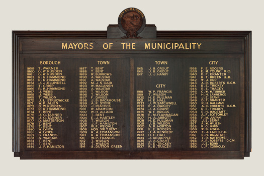 A dark timber rectangular honour board with names listed in gold lettering of the Mayors of Brighton from 1859 to 1965. At the top of the honour board is a round carved crest of the City of Brighton