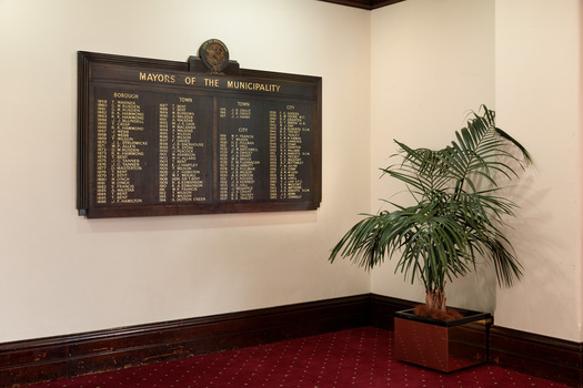 Photograph of a dark timber rectangular honour board with names listed in gold lettering of the Mayors of Brighton. The board is hung on a white wall in the corner of a room with a plant nearby.