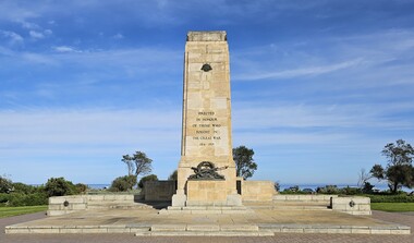 Large stone cenotaph with bronze decoration at its base, surrounded by a stone terrace. Behind the memorial are glimpses of the bay.
