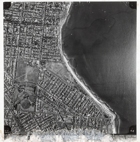 A black and white aerial photograph of a costal suburb with the coast and bay on the right and the streets on the left.