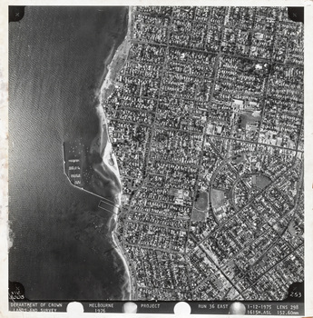 Black and white aerial photograph of a coastal suburb with the bay on the left and house lined streets on the right. Text in the black margin at the bottom of the work