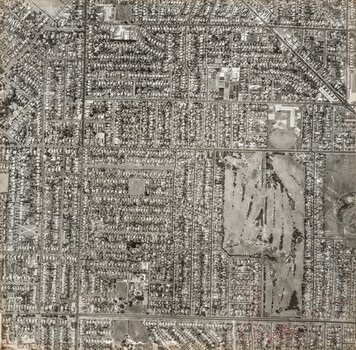 Black and white aerial photograph of streets lined with buildings featuring a large golf course lower right.