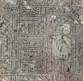 Black and white aerial photograph of streets lined with buildings featuring a large golf course lower right.