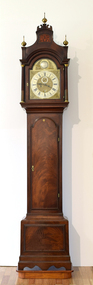 Polished mahogany-cased grandfather clock with three gold spires, chased silver and brass dial and intricately decorated and engraved gold face with Roman numerals. The clock sits on wooden plinth and has a wooden arched door in the centre of its body.
