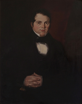 Portrait of seated dark-haired man dressd in a black suit and bowtie with hands clasped on his lap.