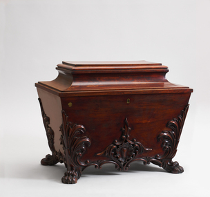 Tapered sides. Scolia shaped lid. Four paw feet. Elaborate carved scrollwork on base and slides. Interior has four lead-lined boxes with hinged lids and holes cut for four magnums of wine.