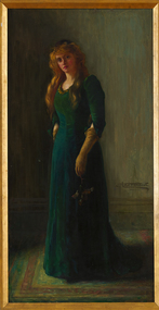 A woman stands in a long green dress with sleeves to the elbow. She wears cream, elbow length gloves and in her left hand holds a sprig of flowers.