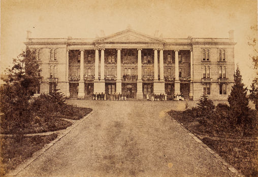 View of a large old building at the end of a road. The building has big columns and a group of people standing at the entrance.