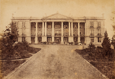 View of a large old building at the end of a road. The building has big columns and a group of people standing at the entrance.