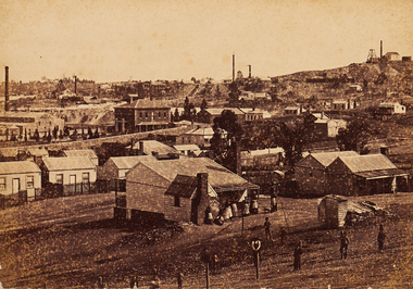 Slightly elevated view of township. Several poppet heads can be seen in the far background. A loose line of figures are present in the foreground.