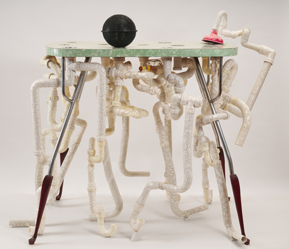 A laminate table top supported by pvc pipes that have been etched and decorated with laser-style cut-out. A black orb with similar cut-outs sits atop the table and a plunger is also present. A whiskey bottle is placed in the tubing just underneath the table top.