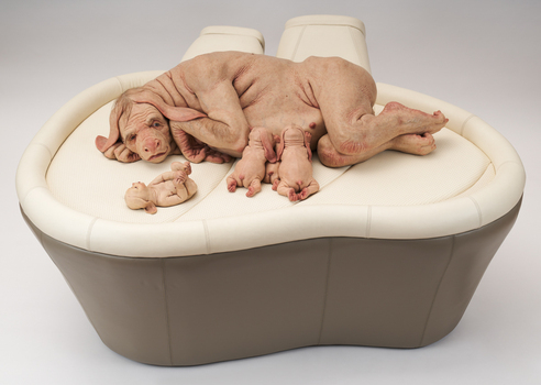 an organic oval shaped plinth upholstered in vinyl supports four alien looking characters. The 'mother' lies in her side while two 'babies' feed at her nipples, one 'baby' lies near the 'mother's' head and rolls on it's back.