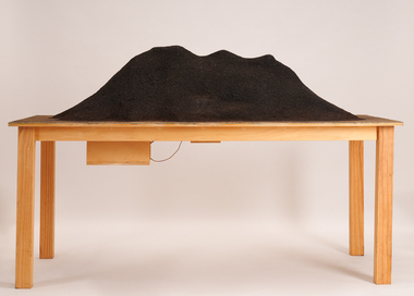 A large wooden table supports a hollow shape resembling a hillside. The hillside is covered in a fine sifting of black sand. A shelf is visible underneath the tabletop where an adapter plug is attached. A cd player can sit in the shelf, and an amp sits inside the hillside.Sound is emitted.